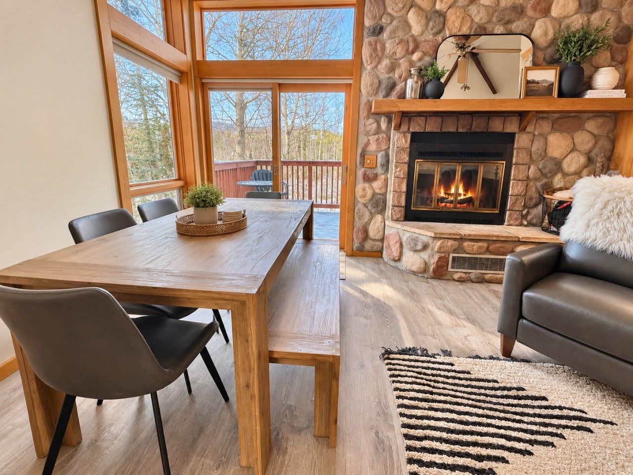 A cozy living room with a stone fireplace, a dining area with a wooden table and chairs, and a view of the outdoors through large windows.