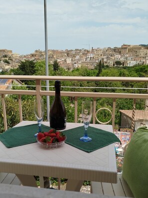 Un tavolo con una bottiglia di vino, due bicchieri e una ciotola di fragole su un balcone affacciato su una cittadina densamente popolata.