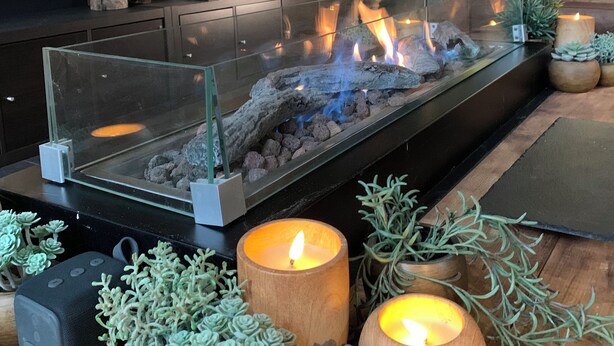 A modern living room with a glass-top fireplace, wooden furniture, and various decorative items including plants and candles.