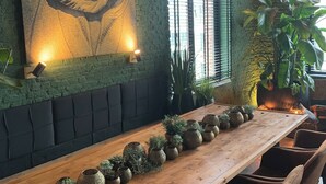 A long wooden table with potted plants, a large painting on the wall, and a brick wall with a window.