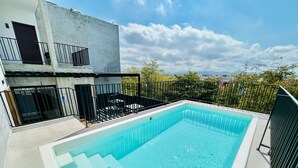 A rectangular swimming pool with a clear blue water, surrounded by a black metal fence, and a white building with balconies in the background. - Cipres Apartments (Mazatlán)