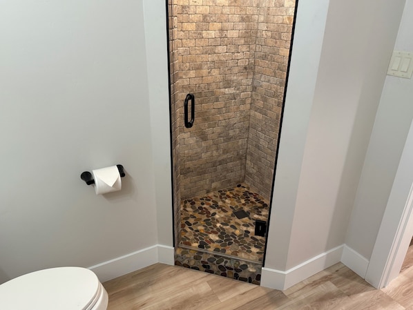 Ein Badezimmer mit Toilette, einer Dusche mit Backsteinwand und Steinboden sowie einem weißen Waschbecken.