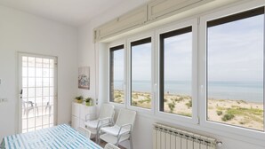 Een kamer met een groot raam dat uitkijkt op een strand, een witte radiator en een tafel met stoelen.