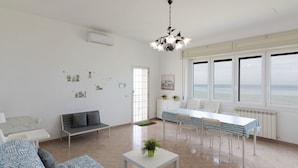 Een ruime woonkamer met een bank, een eettafel met stoelen, een klein witte bijzettafeltje met een plant en uitzicht op zee door grote ramen.
