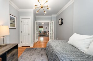 A bedroom with a bed, a nightstand, a lamp, a clock, and a door leading to another room.