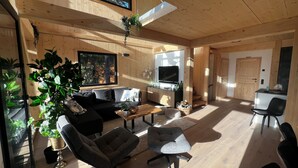 Ein modernes Wohnzimmer mit einer Couch, Sesseln, einem Couchtisch und einem Fernseher. Pflanzen, ein Fenster und eine Tür sind zu sehen.