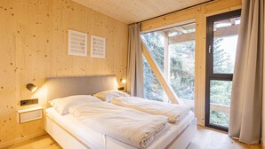 Un dormitorio de cabaña de madera con dos camas, una mesita de noche, una lámpara fijada a la pared y un ventanal con cortinas.