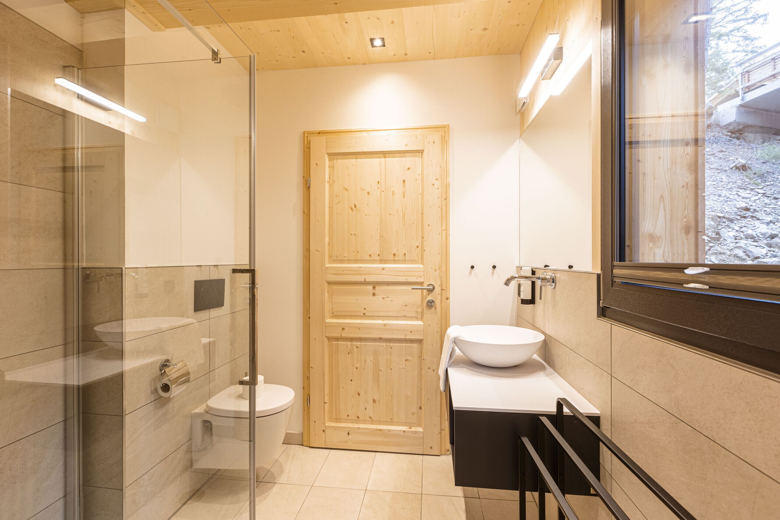 A modern bathroom with a wooden door, a glass shower enclosure, a white sink, and a large mirror.