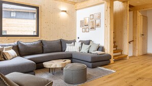 Una sala de estar moderna con un sofá seccional, una mesa de centro redonda y un pequeño otoman. Hay una escalera de madera que conduce a otra habitación, y una pared decorada con cuadros.