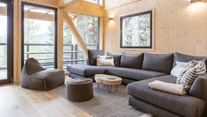 Interior de una cabaña de troncos moderna con sofá modular, sillón de peluche, reposapiés y una mesita redonda. Amplias ventanas ofrecen vistas del bosque circundante.