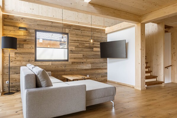 A modern living room with a grey sectional sofa, a flat-screen TV mounted on a wooden wall, a wooden floor, and a staircase leading to another level. - Superior Chalet # 16 with Sauna & Hot Tub (Turrach)