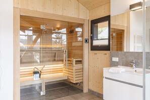 A modern bathroom with a glass-enclosed sauna, wooden benches, a white sink, and a large mirror. - Superior Chalet # 16 with Sauna & Hot Tub (Turrach)