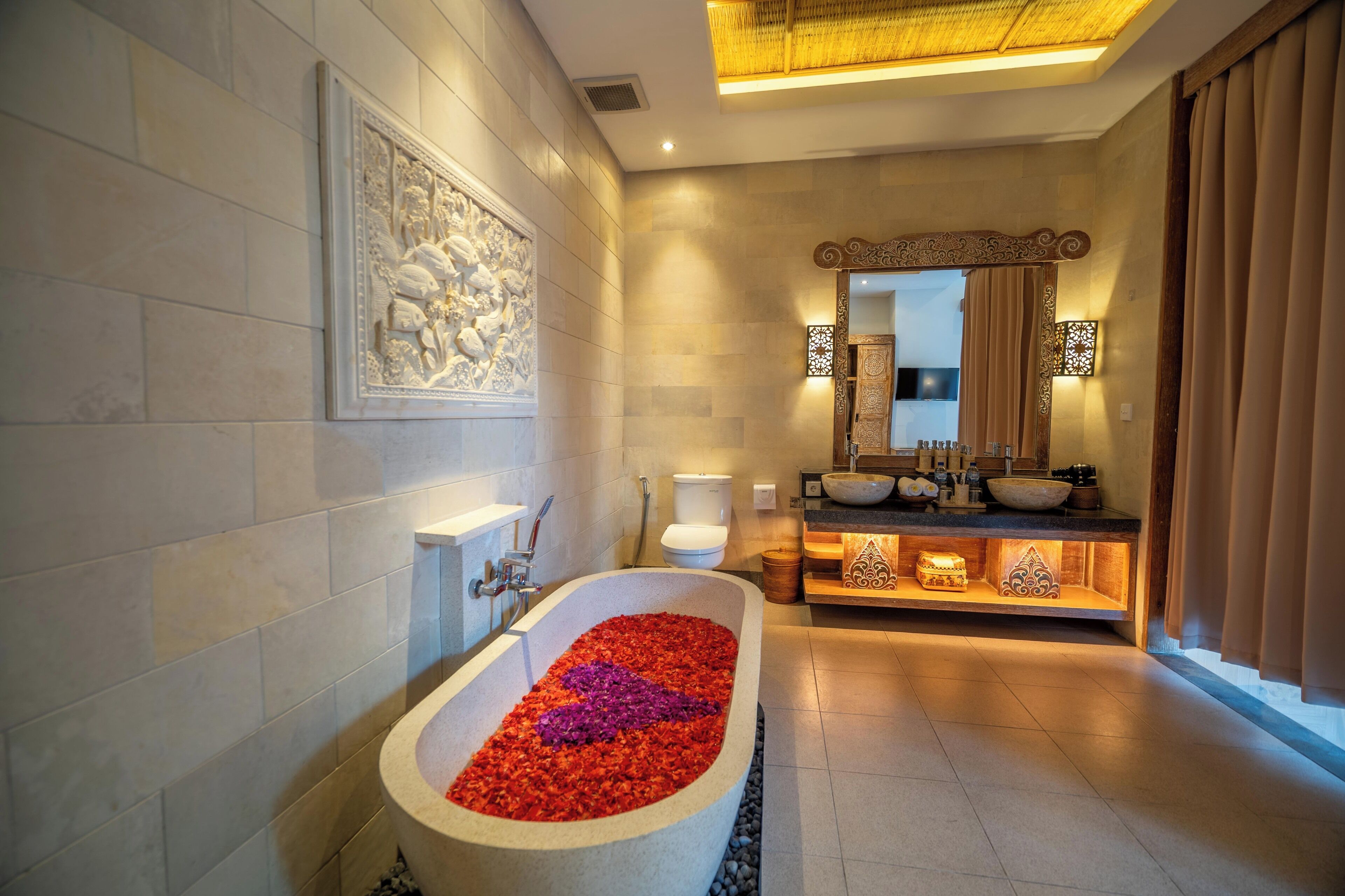 Un baño moderno con una bañera independiente llena de pétalos de rosa, un espejo grande y un tocador con lavabo.