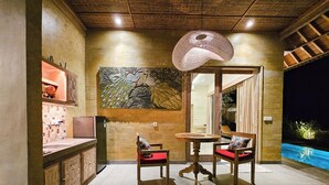Un comedor con una mesa redonda y dos sillas, un adorno en la pared y vistas a una piscina al aire libre de noche.
