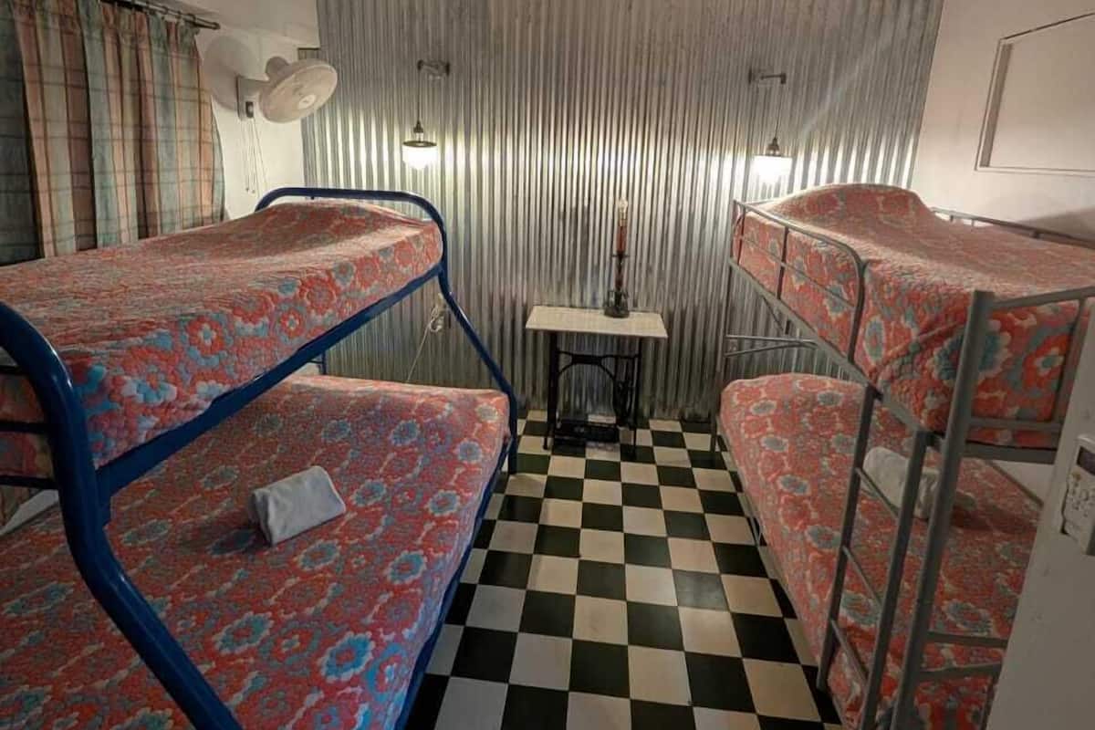 A dormitory room with bunk beds, a checkered floor, a small table, and a wall-mounted fan.