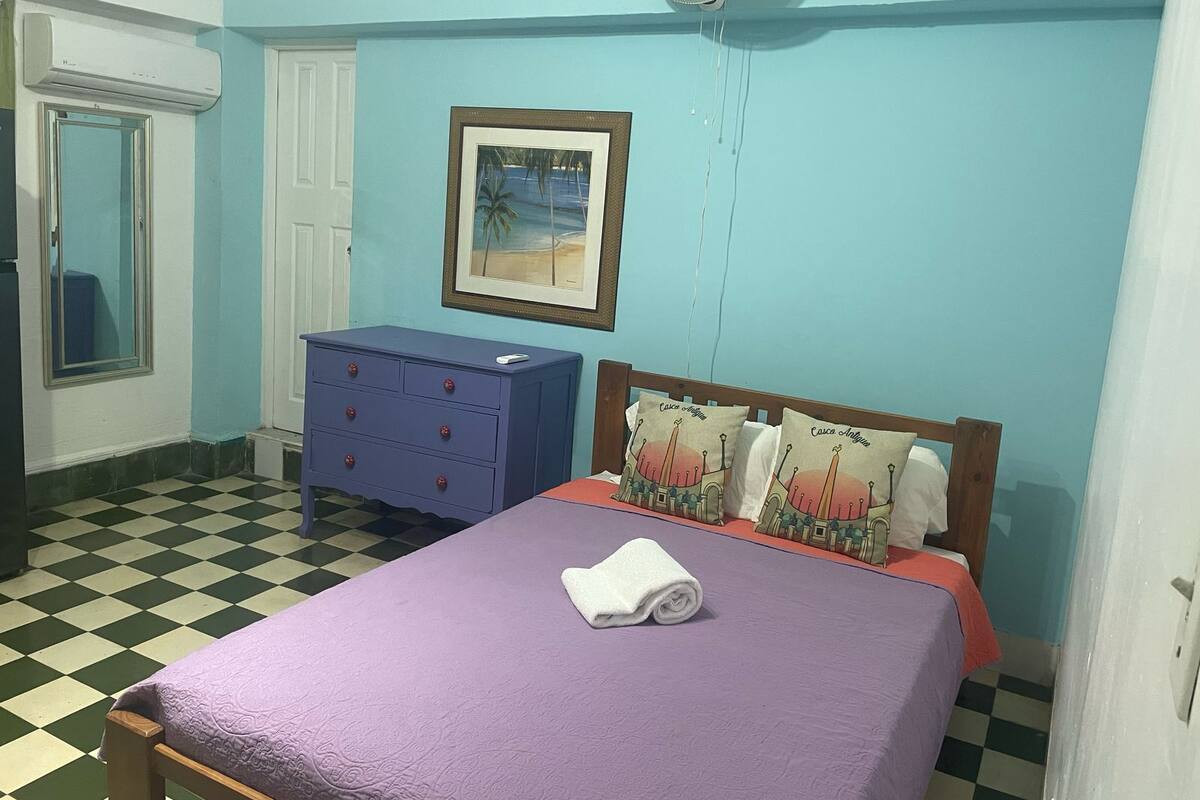 A room with a checkered floor, a bed with a purple bedspread and pillows, a blue dresser, a framed picture on the wall, and a white air conditioning unit.