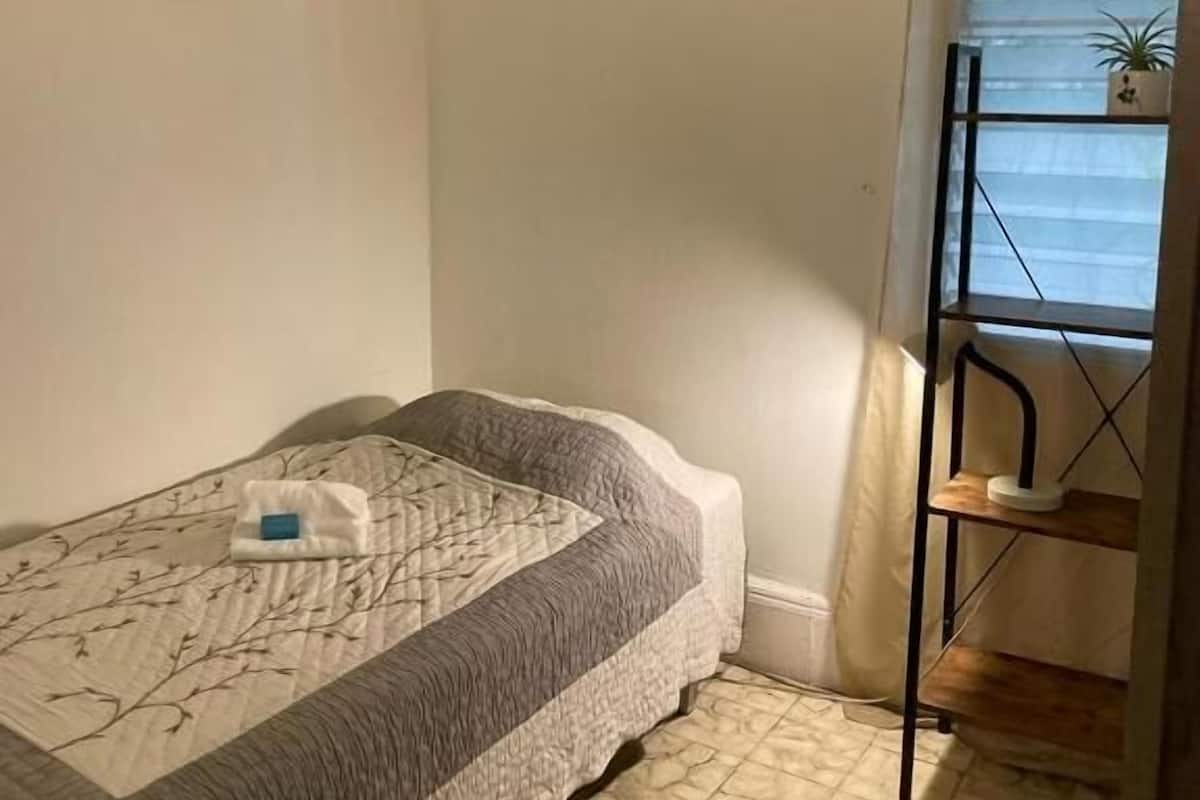 A small, single-bed room with a quilted bedspread, an air conditioning unit mounted on the wall, a window with a curtain, and a small shelf with a plant.