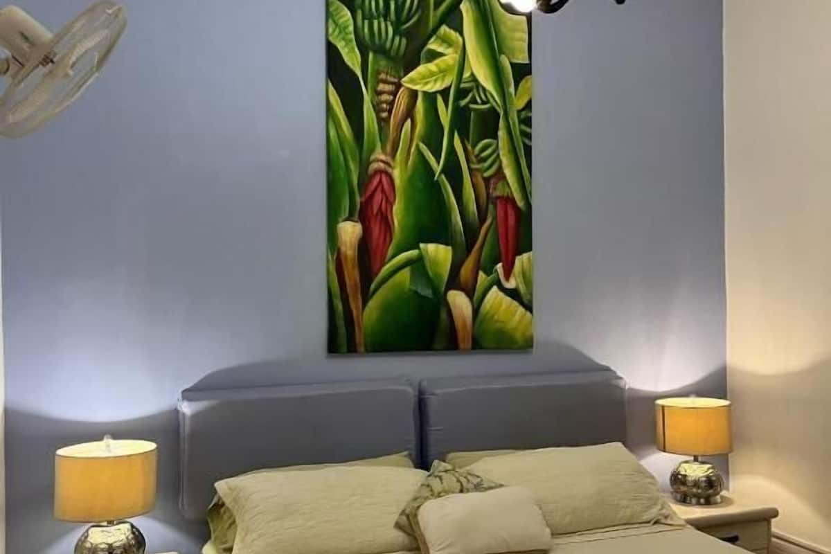 A bedroom with a bed, two bedside tables with lamps, a ceiling fan, and a large abstract painting on the wall.