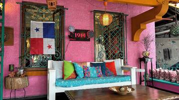 Una habitación vibrante con una pared texturizada de color rosa, un sofá blanco con cojines de colores, una mesa de madera y una ventana con reja de metal.