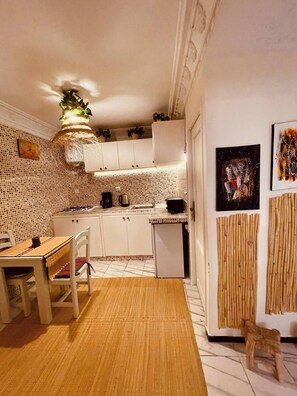 Una cocina con armarios blancos, una mesa de comedor con sillas, un refrigerador pequeño y una planta decorativa colgando del techo.