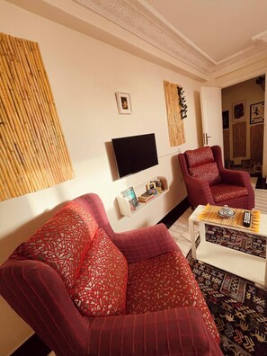 Una sala de estar con dos sofás rojos de estampado, un televisor de pantalla plana, una pequeña estantería y un panel decorativo en la pared.