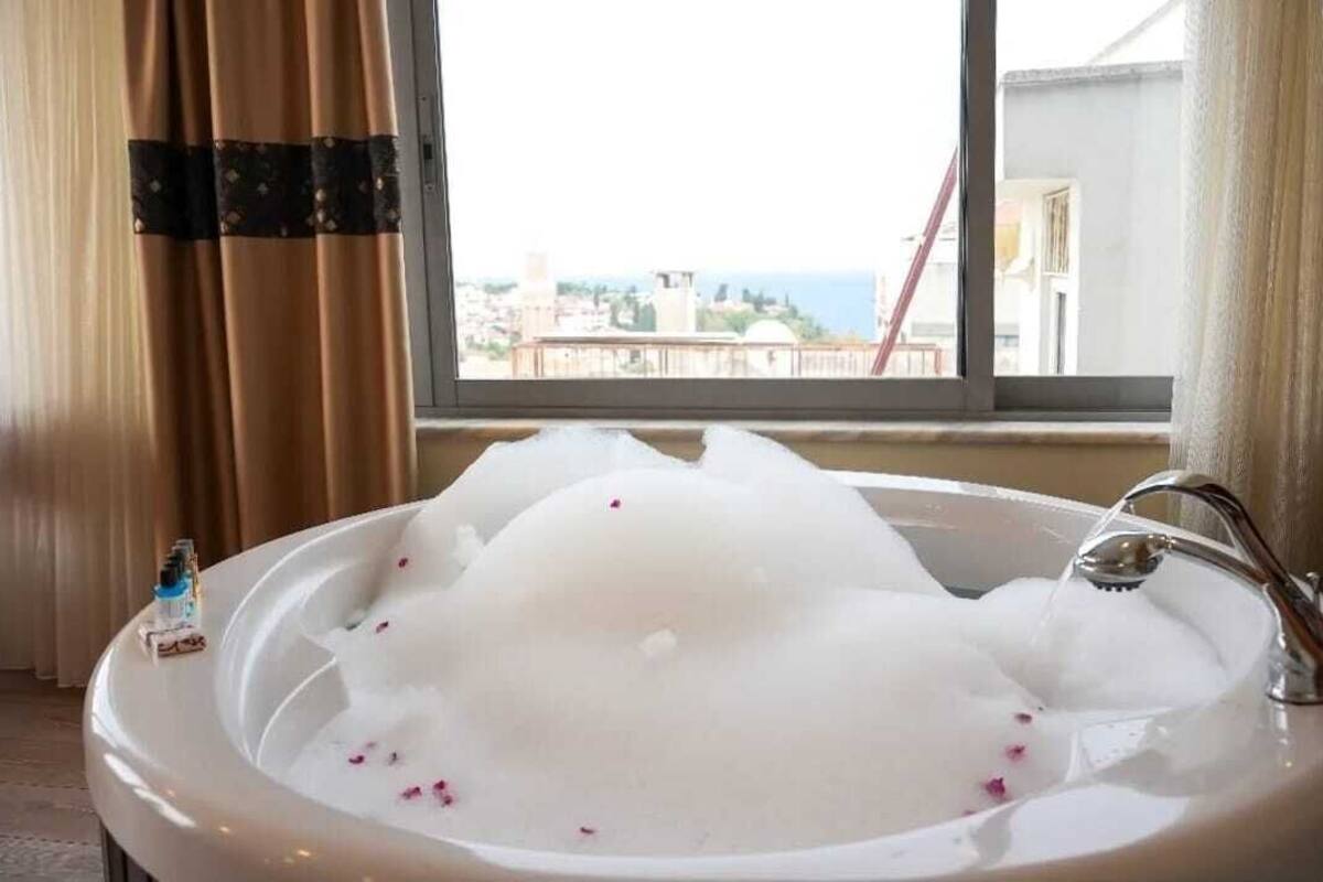 Una bañera llena de burbujas y pétalos de rosa, con vista a un paisaje urbano a través de una ventana.