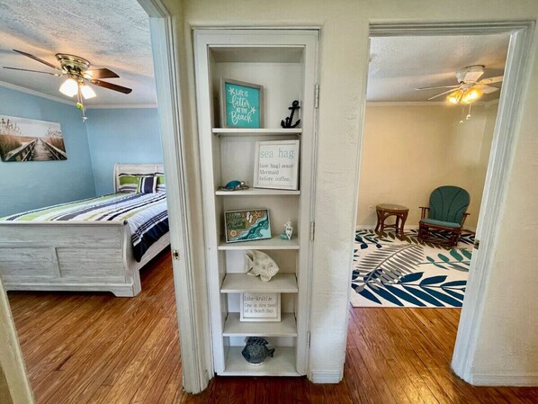 A bedroom with a bed, a bookshelf, and a view into another room with a chair and a patterned rug.