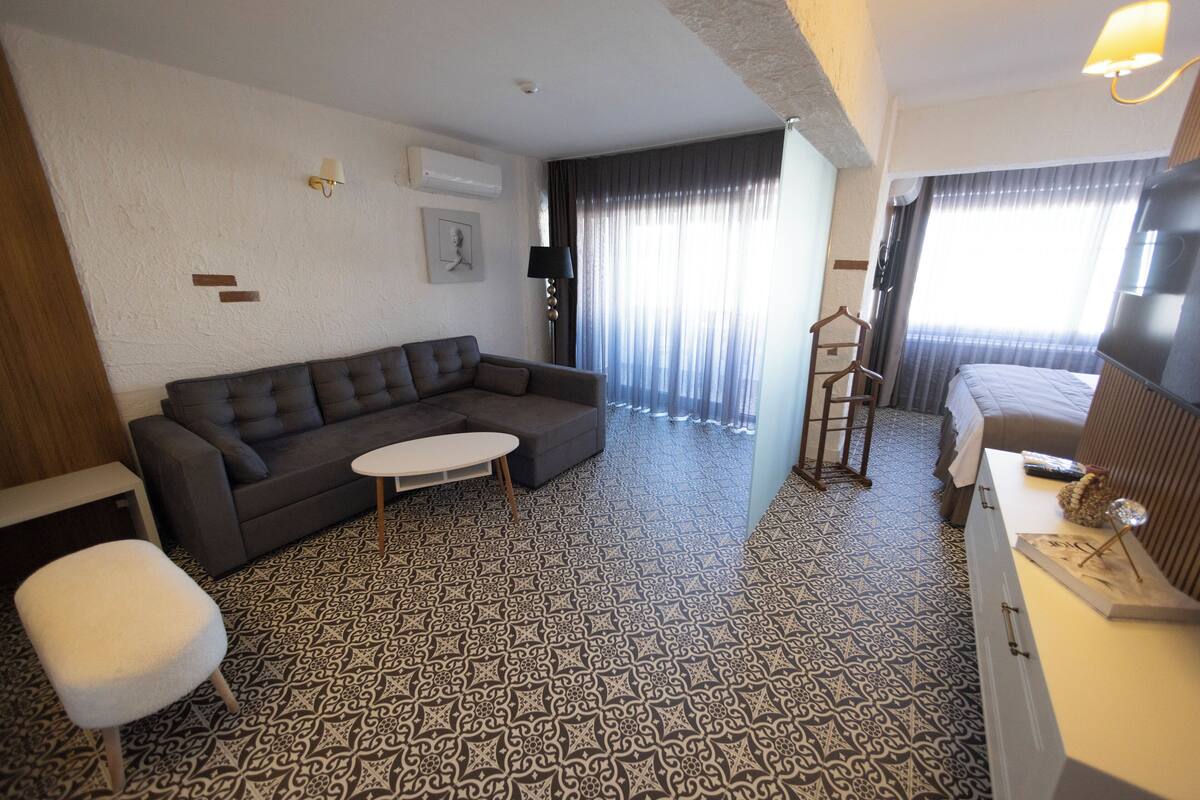 Habitación de hotel con un sofá gris, una mesita redonda pequeña, una silla blanca, una cama con televisor y baldosas de suelo con diseño.