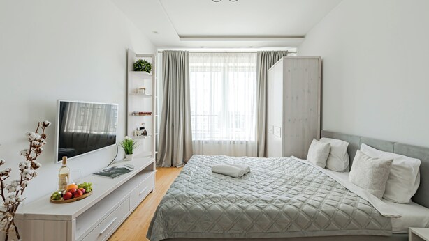 Quarto moderno com uma cama grande, uma televisão sobre um console branco, um guarda-roupa e uma janela com cortinas.