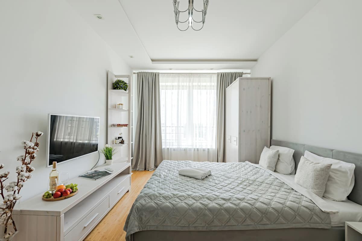 A modern bedroom with a large bed, a TV on a white console, a wardrobe, and a window with curtains.