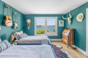 A bedroom with two beds, a dresser, a guitar, a clock, and a large window with a view of the water. - Stunning Waterfront Home with Hot Tub, Deck, Fire Pits, Office & More (Dennis)