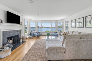 A living room with a fireplace, a large window overlooking a body of water, and a grey sofa. - Stunning Waterfront Home with Hot Tub, Deck, Fire Pits, Office & More (Dennis)