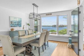 A dining area with a large table, chairs, and a view of a lake through large windows. - Stunning Waterfront Home with Hot Tub, Deck, Fire Pits, Office & More (Dennis)