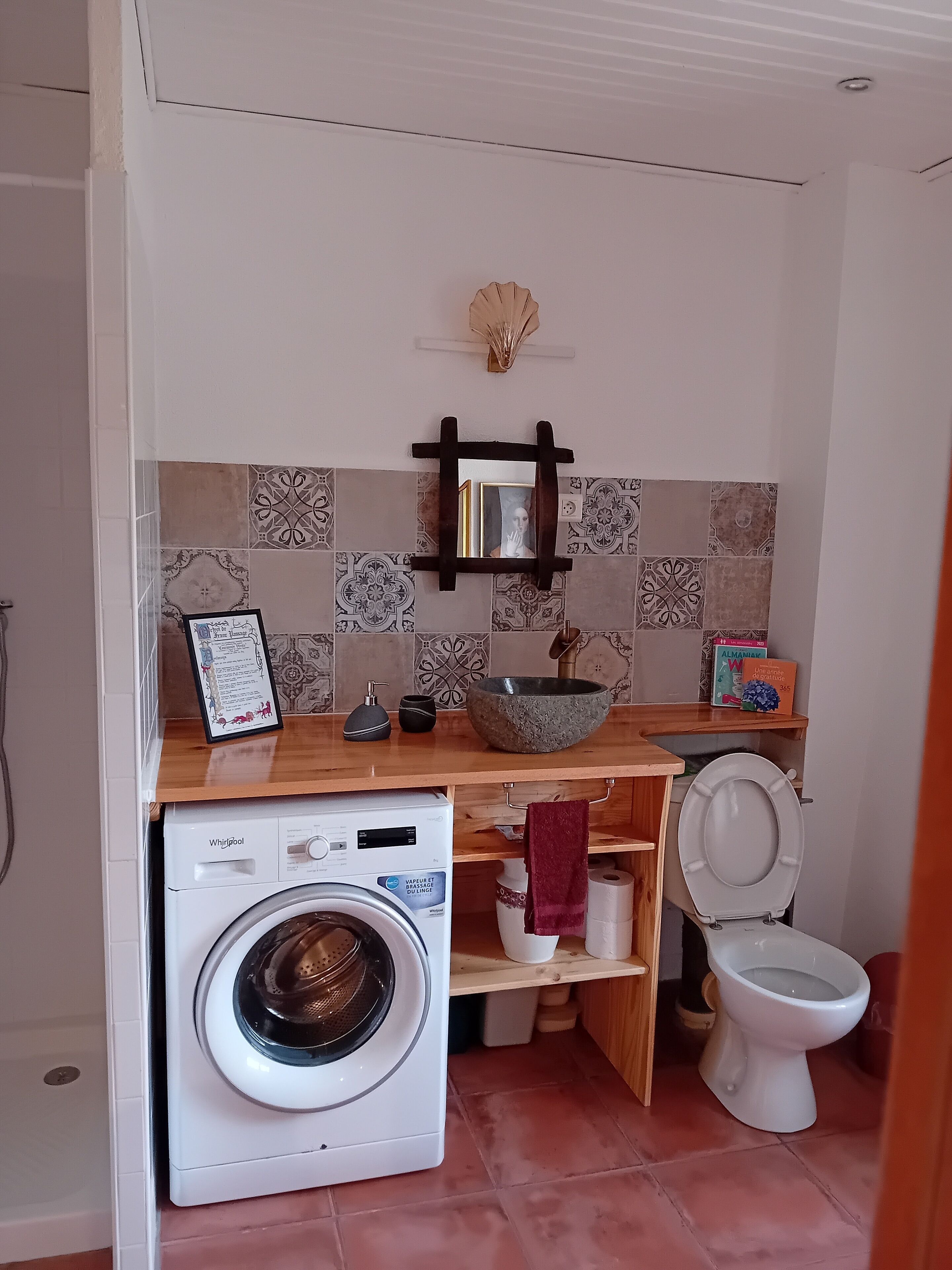 Une petite salle de bain équipée d’un lave-linge, de toilettes et d’une étagère où sont rangés les produits de toilette.