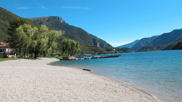 Una spiaggia di ciottoli con una fila di alberi, un pontile con barche e montagne sullo sfondo.