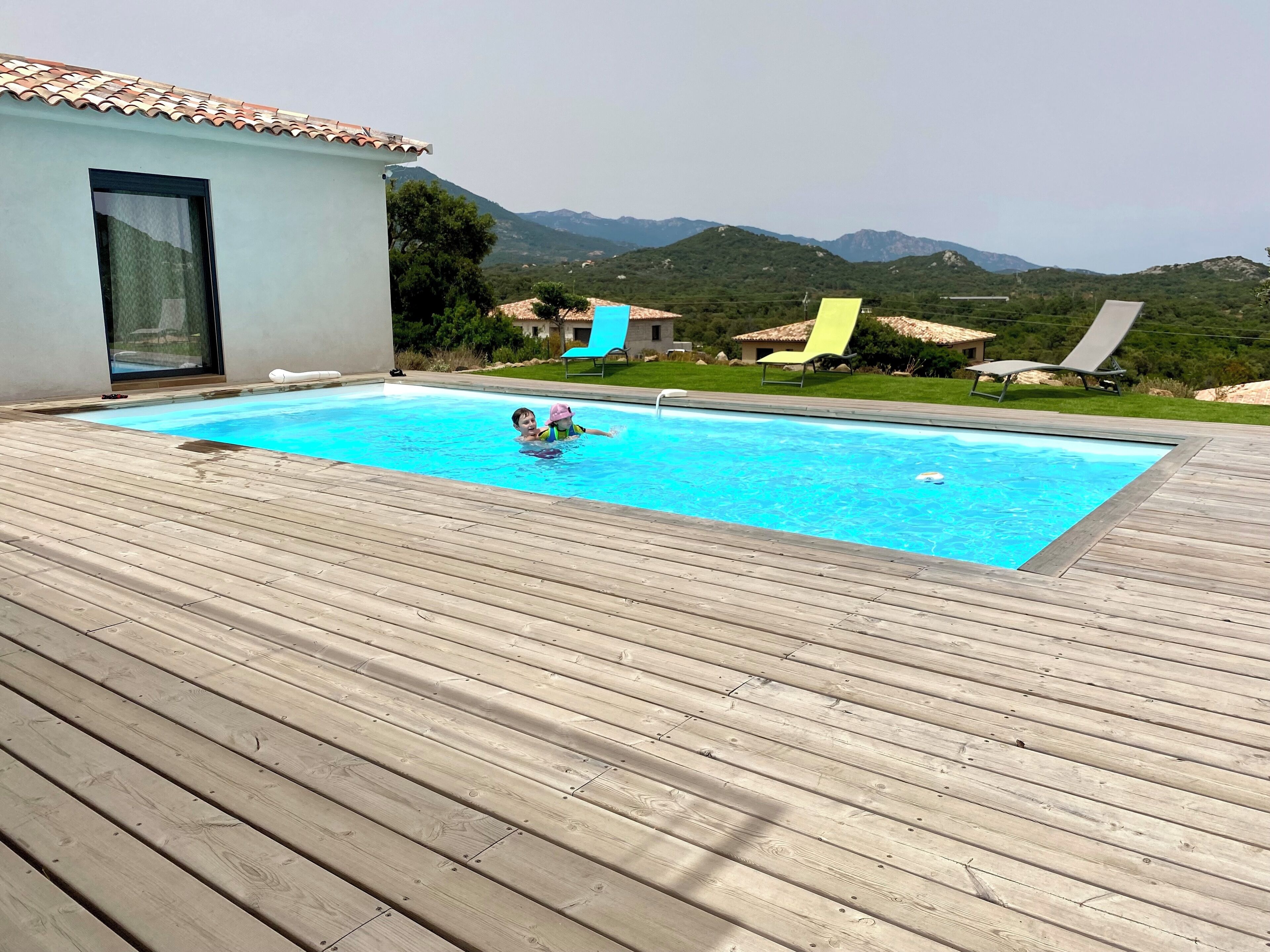 A swimming pool with a wooden deck, surrounded by mountains and a small building with a tiled roof.