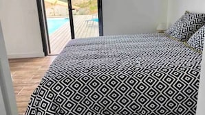 A bedroom with a patterned bedspread, a view of a pool and hills through large windows. - Villa de Standing Avec T2 Indépendant vue Panoramique (Sotta)