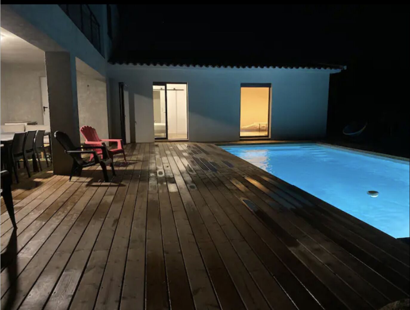 A wooden deck with a pool, a dining area with chairs, and a door leading to a bright interior.