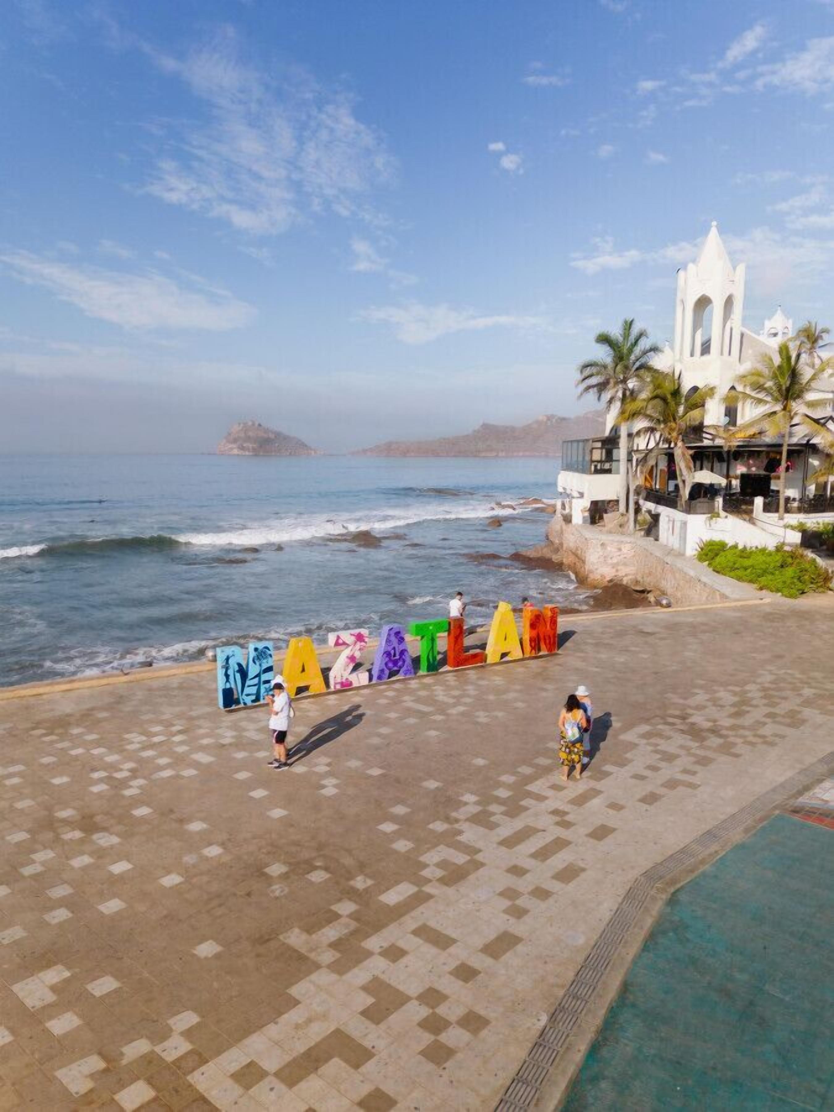 Un hotel costero con vistas al mar, una iglesia blanca y letras coloridas que forman la palabra 'ATLANTIS' en el suelo.