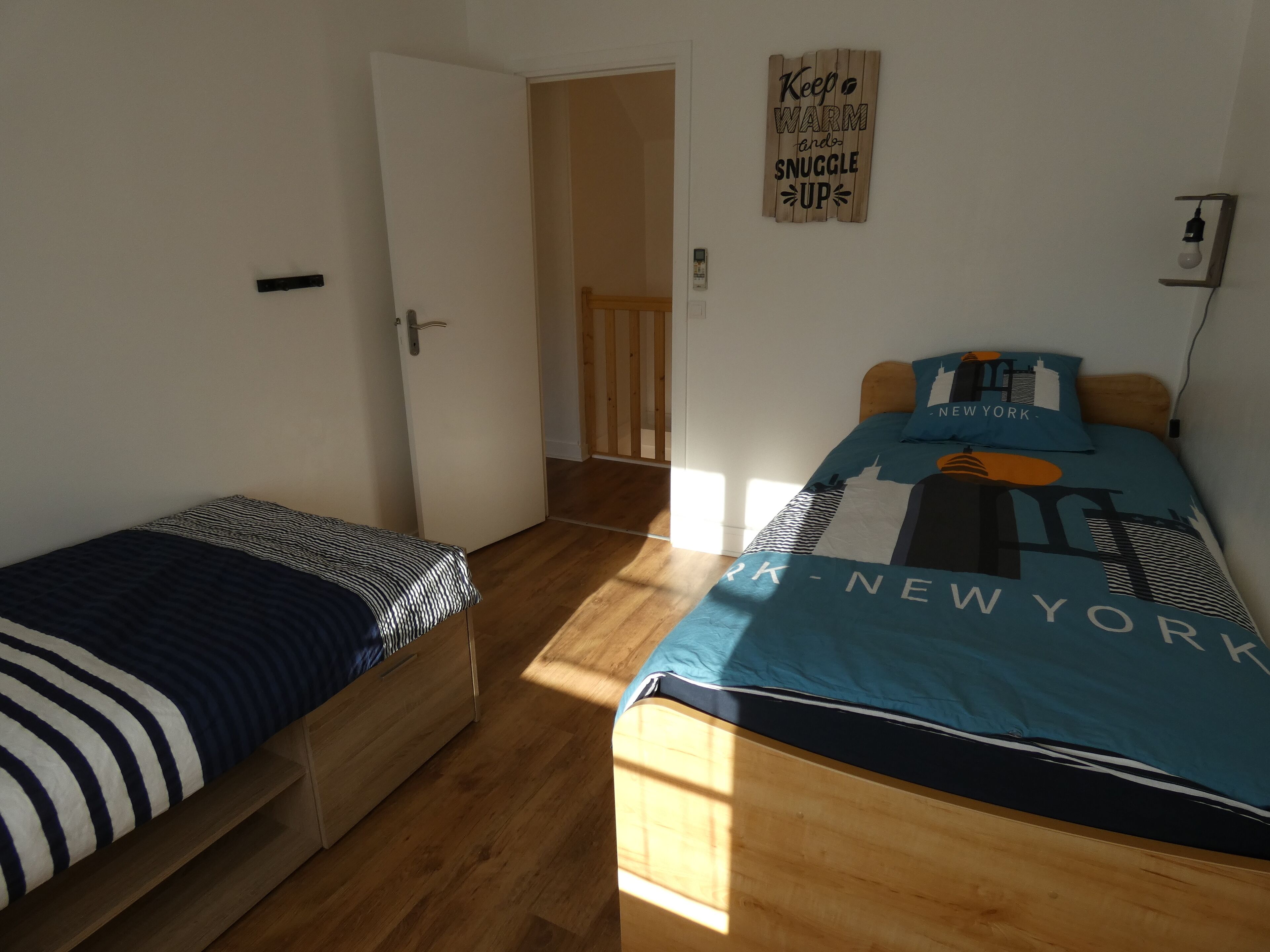 A small, well-lit room with two beds, wooden flooring, and a wall-mounted light fixture.