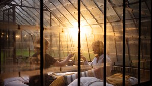 A greenhouse with a glass ceiling, allowing natural light to enter. Inside, there are beds with pillows and blankets, and a person is visible in the background. - La Petite For t Antique Countryside Greenhouse (Asse)