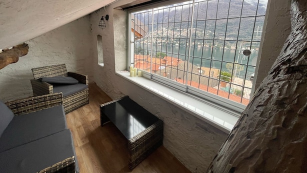 A room with a large window overlooking a body of water and a cityscape. There are wicker chairs and a small table. - Timco apartment with lake view (Torno)