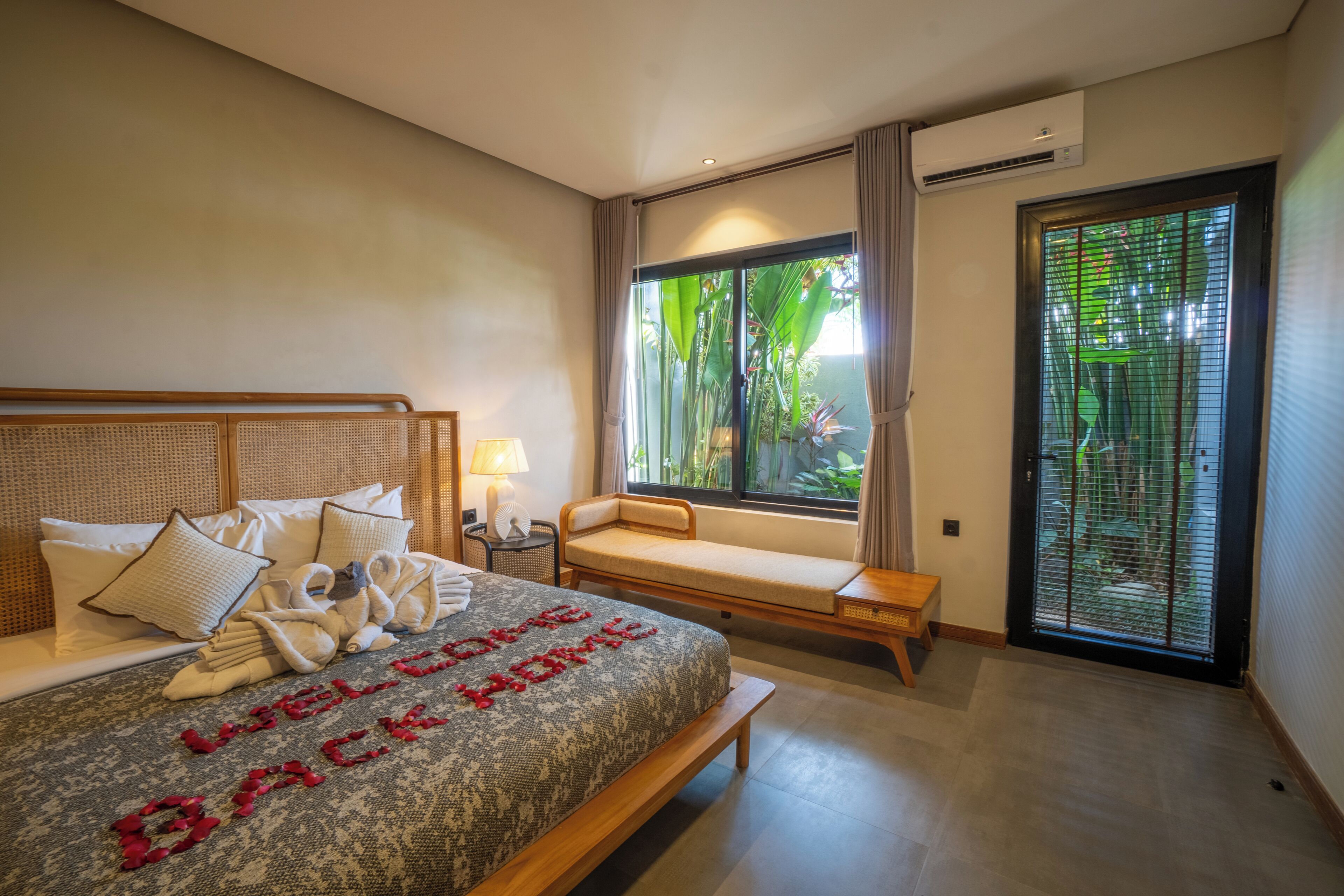 A hotel room with a bed, a bench, a window with a view of greenery, and an air conditioning unit.