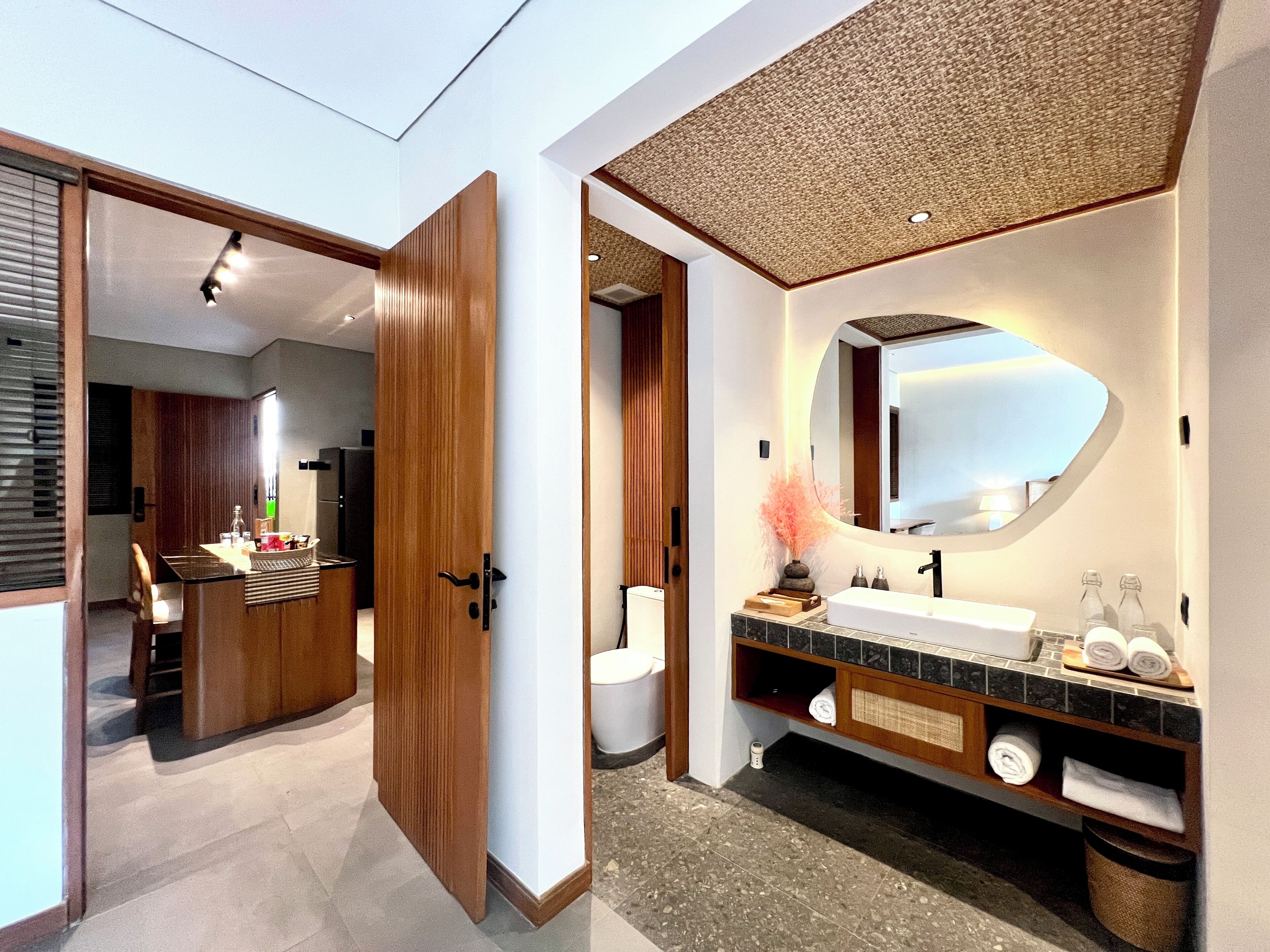 Ein modernes Badezimmer mit einem großen Spiegel, einem Waschbecken und einer Toilette. Eine Holztür führt in einen anderen Raum.
