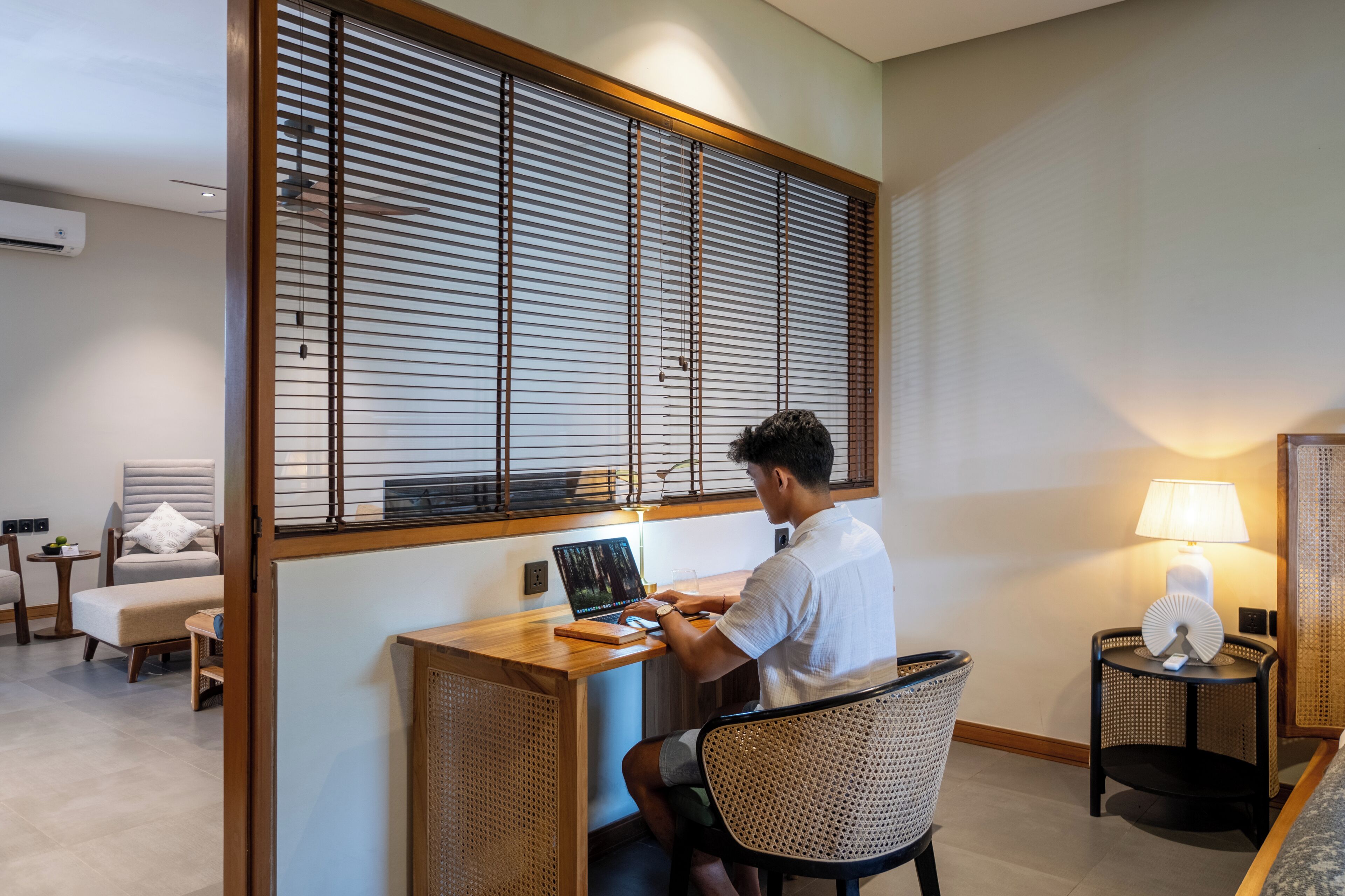 A man working at a wooden desk in a modern room with a large window, a lamp, and a chair.