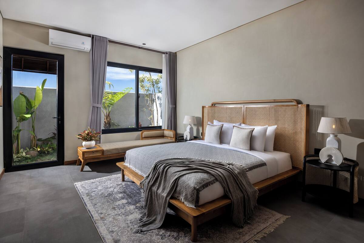 A modern bedroom with a large bed, bedside tables, a window with curtains, and a view of greenery outside.