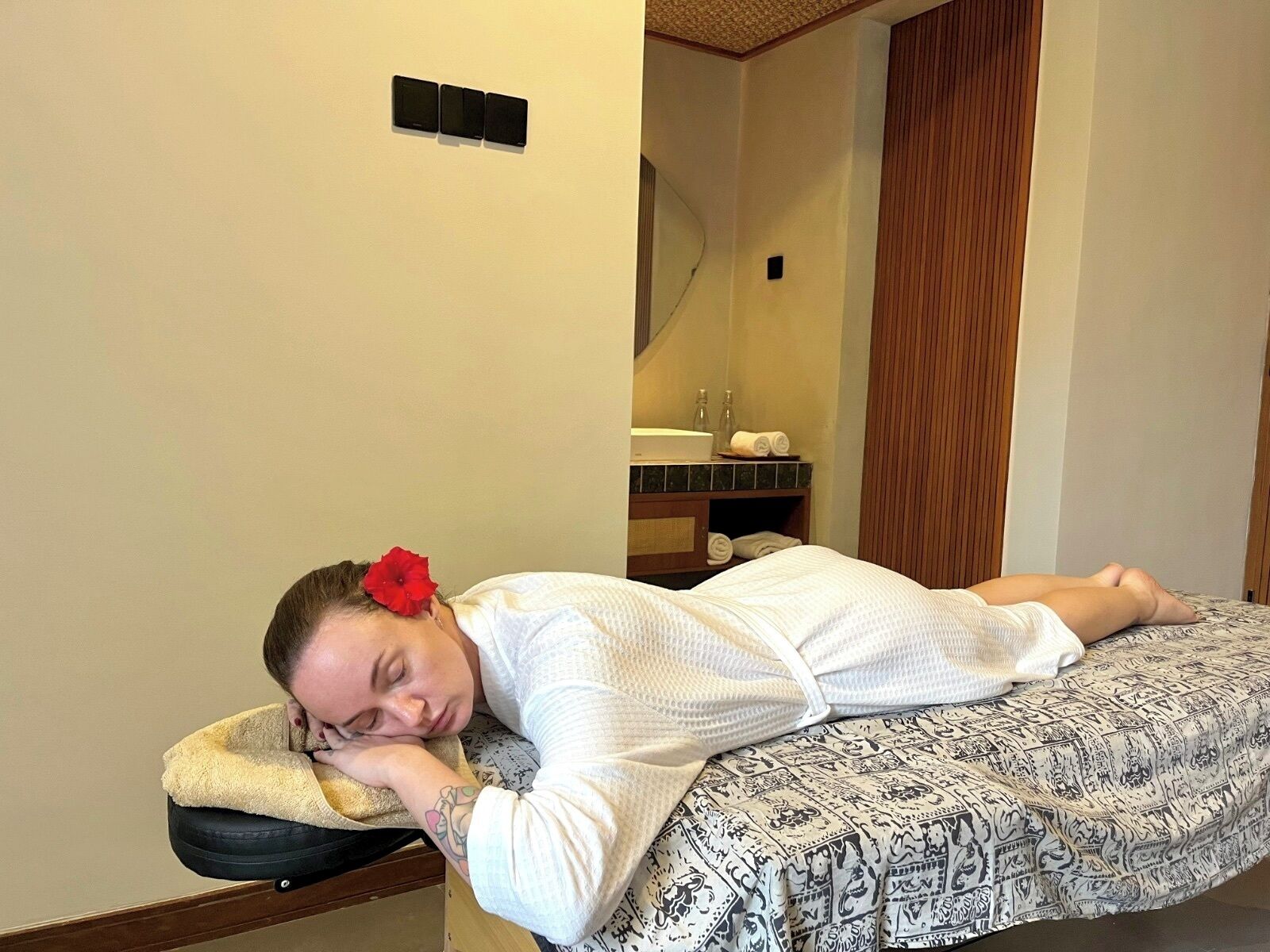 A person lying on a bed in a room with a wooden floor, a mirror, and a sink with towels.