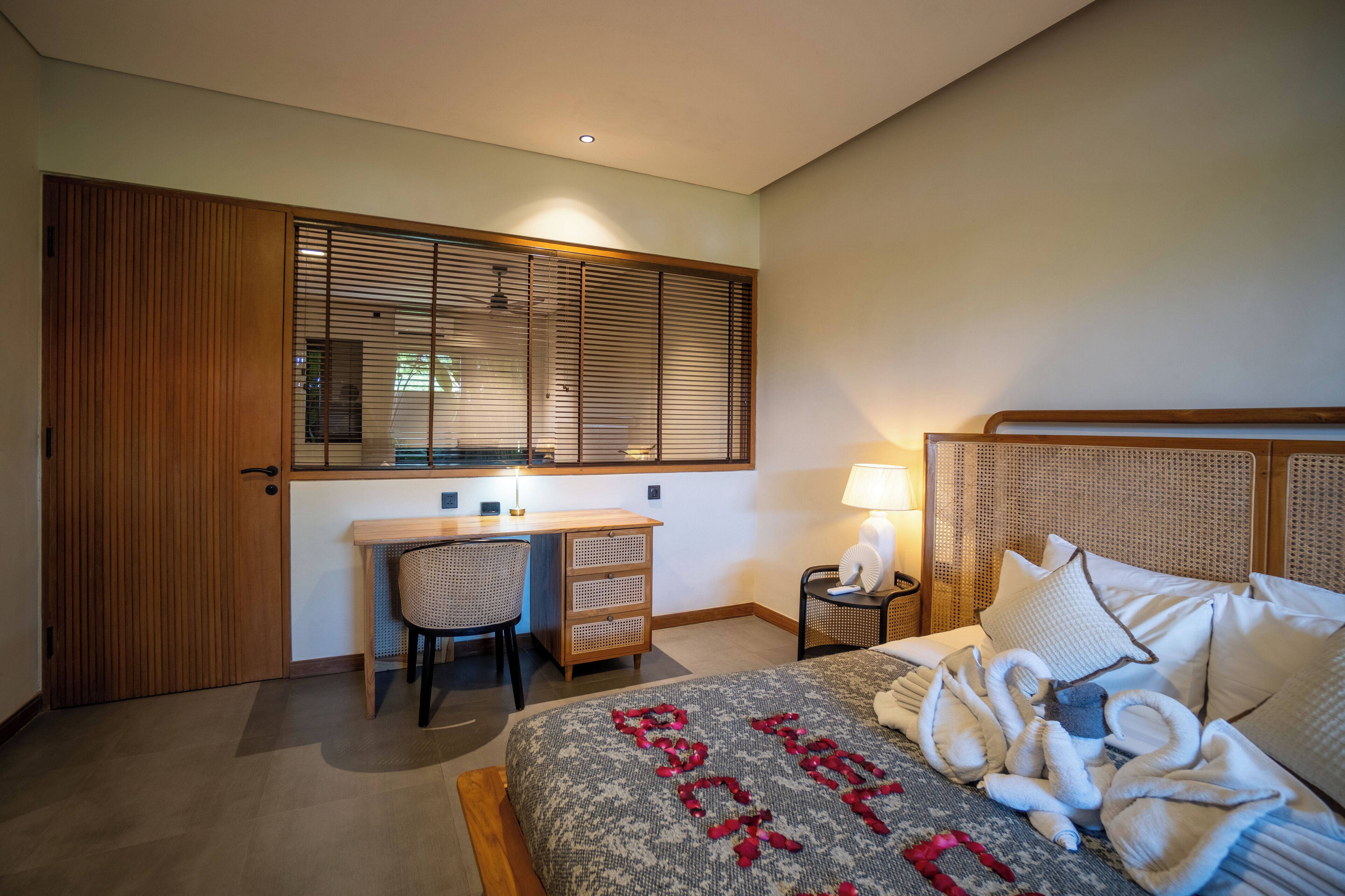 A hotel room with a bed, a desk, a chair, a bedside table with a lamp, and a large window with blinds.