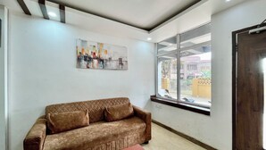 A room with a brown sofa, a wooden coffee table, a ceiling fan, a window with a view of buildings, and a painting on the wall. - Hotel Subham Beach Inn - near Sea Beach (Puri)