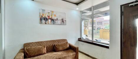 A room with a brown sofa, a wooden coffee table, a ceiling fan, a window with a view of buildings, and a painting on the wall.